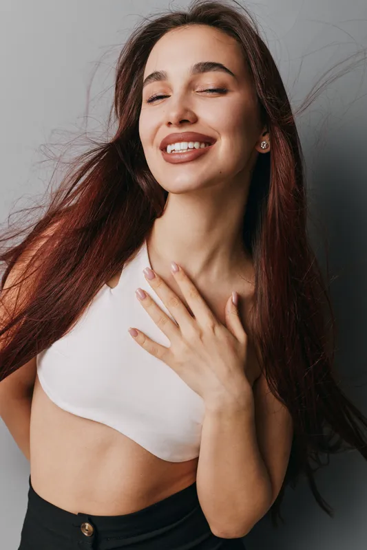 Woman smiling with closed eyes and hand on chest wearing white crop top and black pants standing against gray wall. Long hair and natural makeup with joyful expression.