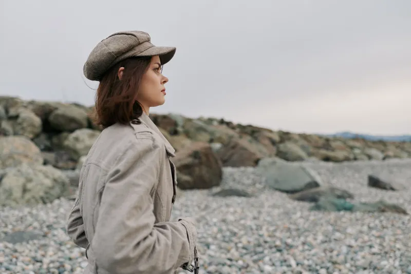 Young woman in beige coat and hat stands on rocky beach looking into distance. Overcast sky creates moody atmosphere. Concept of contemplation, nature, and autumn fashion outdoors.