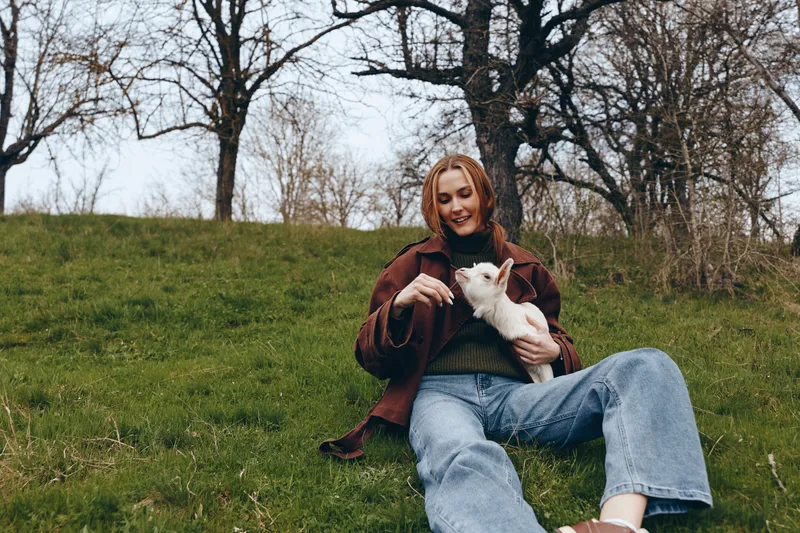 A young woman in a brown jacket and blue jeans sits on a grassy hill while gently holding and feeding a white baby goat in a natural outdoor setting during early spring.