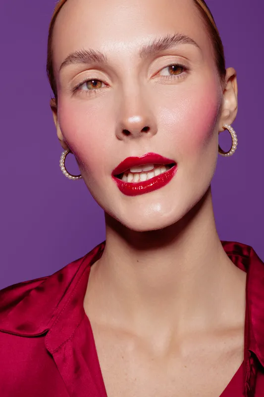Close-up portrait of a woman with glossy red lips and hoop earrings wearing a red blouse against purple background. Concept of beauty, makeup, fashion, and elegance.