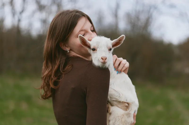 Woman in a brown sweater embraces a friendly goat kid in a rural field. Soft natural light creates a warm, intimate lifestyle moment, highlighting tenderness, care, and outdoor happiness in a