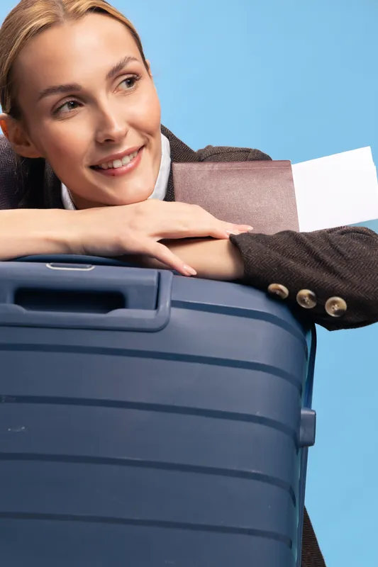 Smiling woman leans on blue suitcase holding a boarding pass, ready for travel. Studio portrait with bright background, concept of vacation, trip, and holiday preparation.