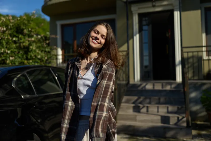 A young woman with curly hair smiles warmly outside a residential building, casually posing in a plaid shirt and denim jeans, as sunlight highlights the welcoming entrance and a relaxed everyday vibe