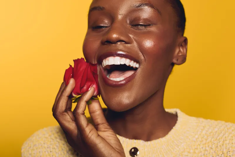 Happy African woman with dark skin laughs and holds red rose near face on yellow background. Concept of joy, beauty, nature, and positive emotions in studio portrait.
