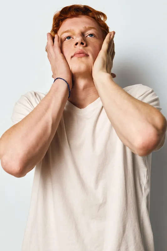 Young man with red hair and fair skin touching face with hands in contemplative pose against white background. Casual style and natural expression in studio portrait.