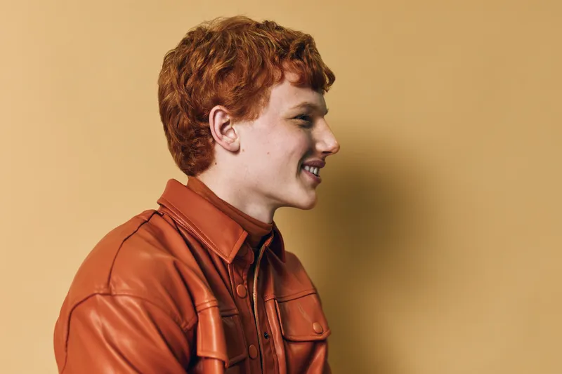 Redhead man with curly hair wearing a brown jacket smiling and looking sideways on beige background in studio portrait with casual style and warm colors.