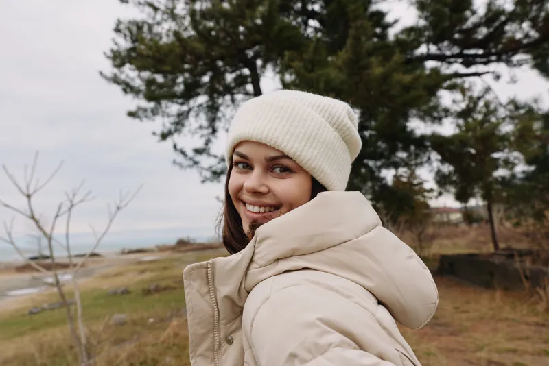Smiling woman outdoors in a winter jacket and knit beanie, landscape with trees and distant coast, portraying warmth, style and fresh air.