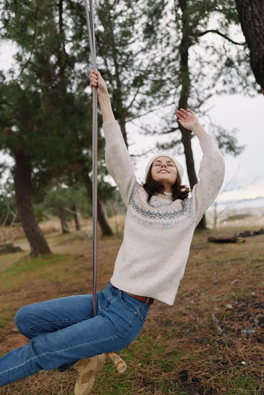 Girl on a swing outdoors, smiling and carefree in a cozy sweater and jeans, among trees and grass, capturing playful nature and youth in a tranquil park setting