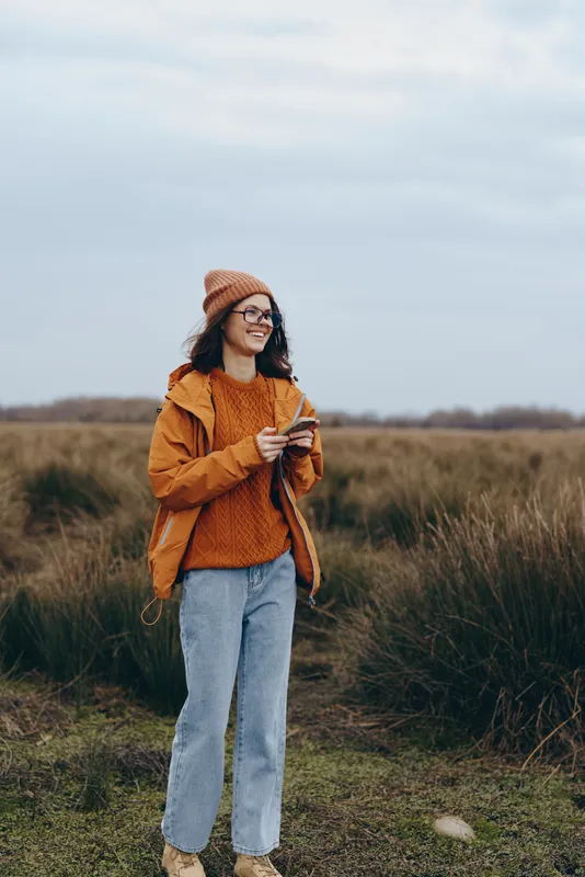 Woman lifestyle outdoors in field at cloudy sky, wearing orange sweater and hat, denim pants, smiling while holding phone, autumn nature scene, soft wind mood during daylight