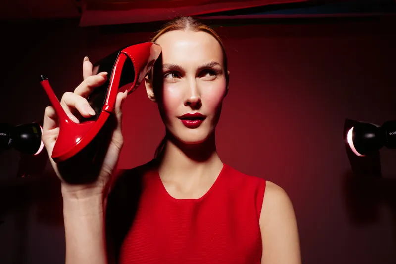 Woman in red dress holds red high heel shoe near face with confident expression. Dark background and dramatic lighting create bold fashion concept.