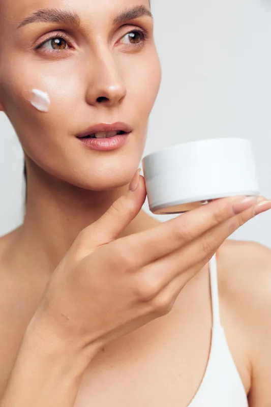 Young woman with clear skin applies moisturizing cream on her cheek while holding a white jar in a minimalistic bright setting, showing a natural skincare routine with a calm expression.
