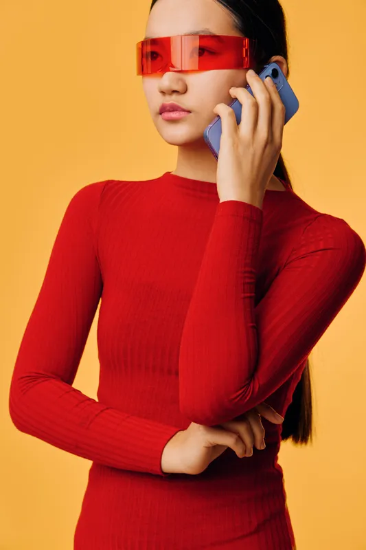 Young woman wearing red dress and futuristic red sunglasses talking on smartphone. Stylish girl with long hair holding phone on yellow background for communication and technology.