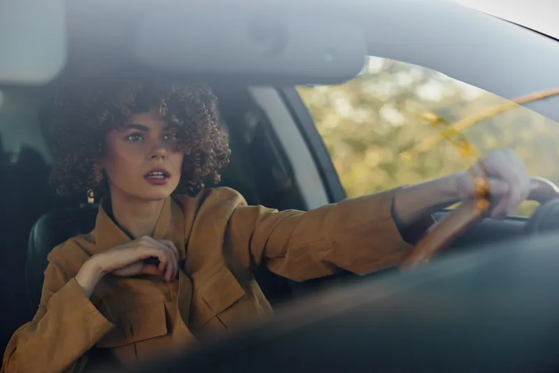 Woman driver in a mustard jacket sits behind the wheel, looking ahead. Close-up on the interior with soft light, retro film color mood, lifestyle and candid moment.