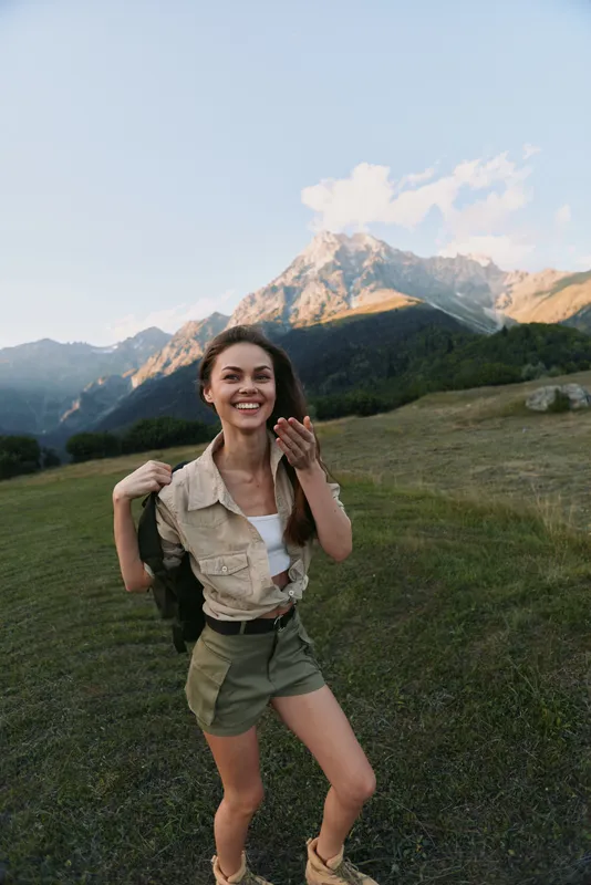A smiling young woman in casual summer clothes stands on a grassy mountain meadow with a scenic peak in the background during daylight. Lifestyle and nature concept.