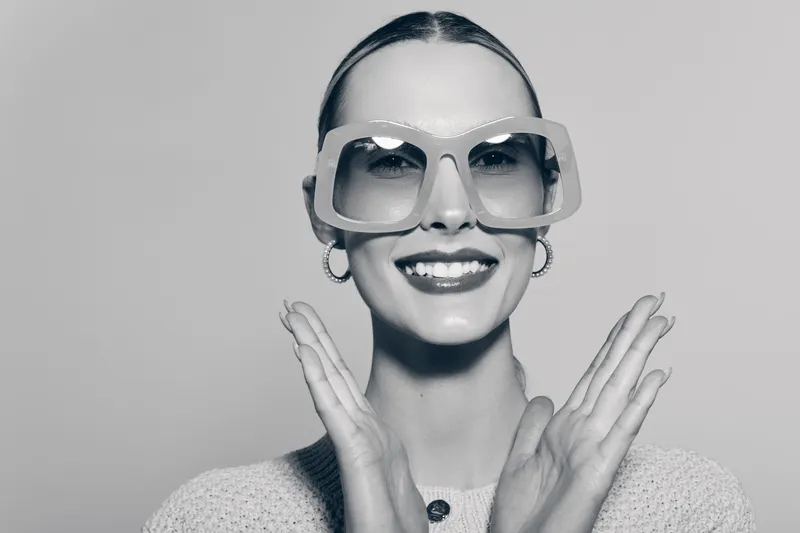 Smiling caucasian woman with pale skin and slicked hair wears large square glasses and knitted sweater. Studio portrait with bright smile and hands near face.