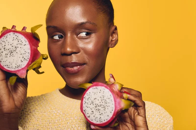 Young African woman with dark skin holds two halves of dragon fruit, smiling slightly, on bright yellow background. Concept of healthy eating, tropical fruit, natural lifestyle, and wellness.