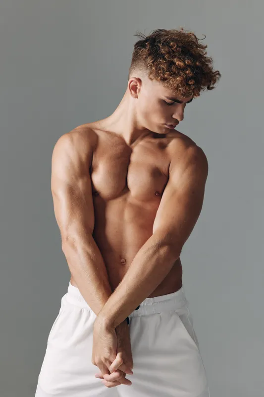 Muscular young man with curly hair posing shirtless in white shorts against gray background, showing toned arms and chest with focused expression.