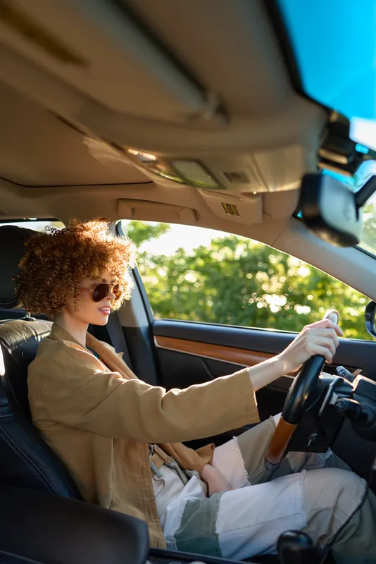 Cheerful woman driving a car outdoors, curly hair, sunglasses, beige coat, sunlit road scene, relaxed posture, natural colors, candid lifestyle moment, vibrant atmosphere