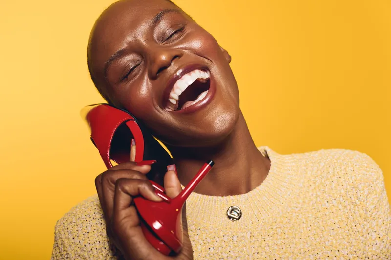 Happy dark skinned African woman with short hair laughing and holding a red high heel shoe near her face on yellow background. Concept of joy, fashion, beauty, and lifestyle.