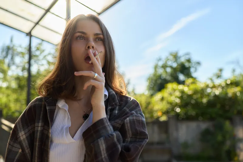 Cheerful woman outdoors, thoughtful moment, finger to lips gesture, sunlit scene, casual plaid shirt, soft breeze, natural colors, leafy background, relaxed pose, candid lifestyle vibe