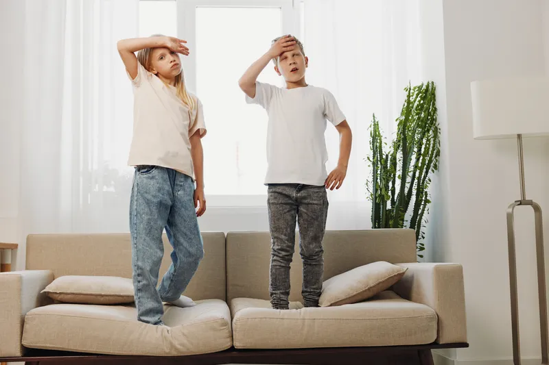 Two children standing on a beige sofa in a bright living room, both with hands on their foreheads as if searching or looking far. Modern home interior with natural light and green plant.