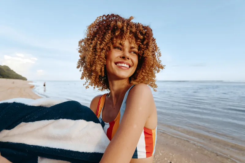 Smiling woman with curly hair on beach by ocean holding blanket, warm sunlight, gentle waves, summer vacation outdoors portrait, joyful relaxed mood, seaside horizon behind, copy space