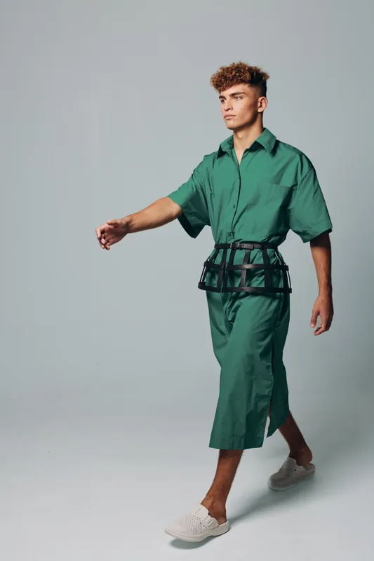 Young man walking in green oversized shirt and matching wide pants with black belt bag and white slip on shoes on gray background in studio. Fashion model with curly hair and casual style.