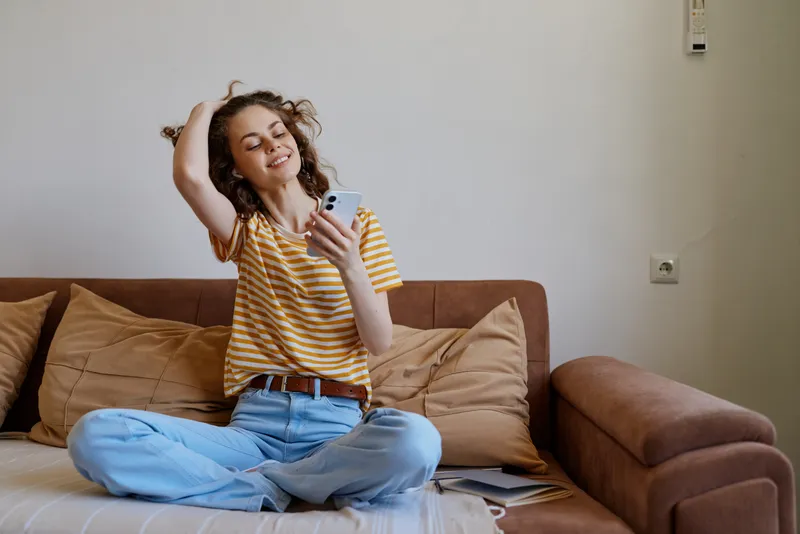 Young woman in a striped tshirt sits on a sofa in a living room, smiling as she looks at her phone, capturing a candid moment of relaxed, everyday happiness at home.