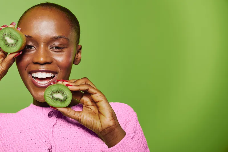 Happy black woman with dark skin holds fresh kiwi slices near face, smiling with teeth. Bright green background and pink sweater create vibrant healthy lifestyle concept with fresh fruit.