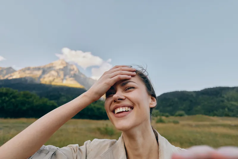 Smiling woman outdoors shielding sun with hand in a sunlit landscape, mountains in background, casual lifestyle moment, natural warmth