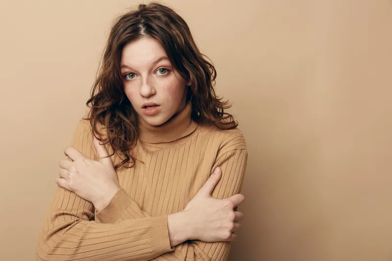 Young woman with curly brown hair wearing beige sweater looks surprised and thoughtful against plain beige background. Concept of emotion, contemplation, and casual fashion portrait.