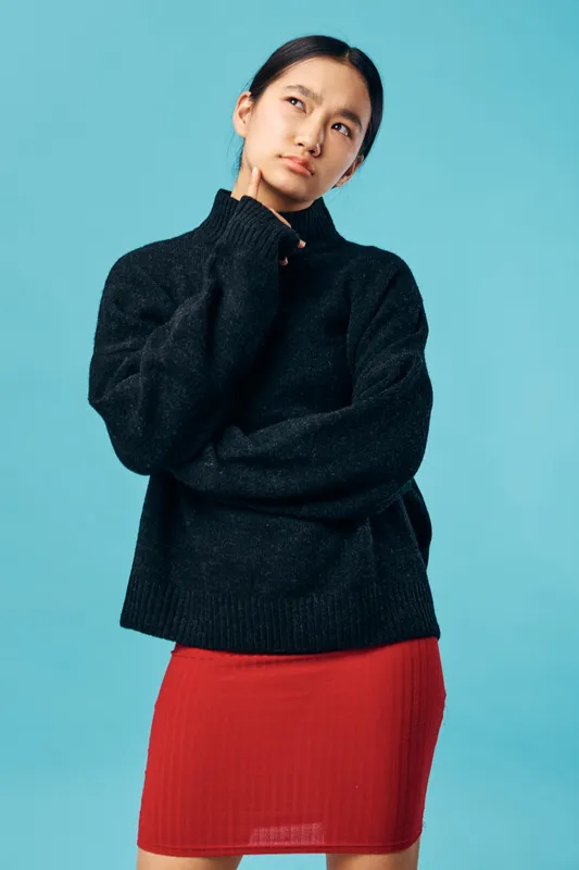 Young woman in black sweater and red skirt standing with thoughtful expression and hand on chin against blue background. Casual fashion and pondering concept with studio lighting.