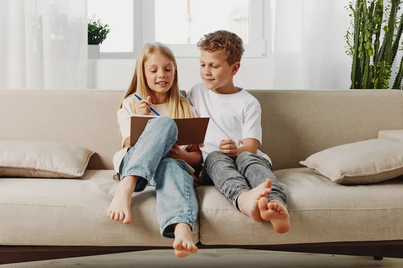 Two barefoot children sitting on a beige sofa, drawing and sharing a creative moment in a bright living room with plants and cushions.