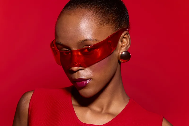 African American woman with dark skin wearing red sunglasses and red sleeveless top poses against red background. Stylish portrait with bold colors and modern fashion concept.