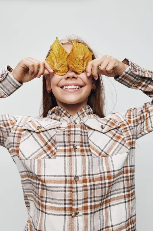 Girl smiling and holding two autumn leaves in front of her eyes wearing plaid shirt on white background. Child playing with nature elements in fall season and having fun indoors.