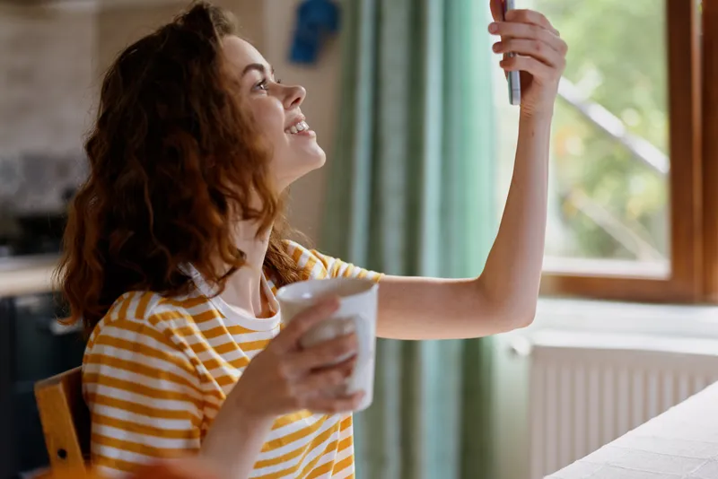 Young woman in a striped yellow shirt takes a carefree selfie indoors near a sunlit window, smiling warmly while holding a white cup and enjoying a casual moment at home.