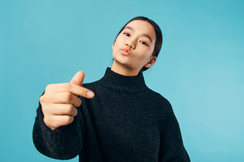 Woman in black sweater making finger heart gesture with lips puckered against blue background. Young female showing love symbol, casual portrait with confident expression and studio lighting.