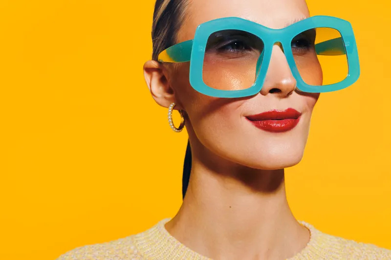 Stylish woman with large turquoise sunglasses and red lipstick poses against bright yellow background. Concept of fashion, summer, beauty, and confident attitude.