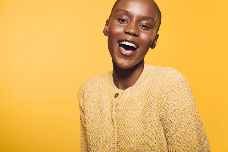Happy African woman with dark skin laughing in yellow knitted sweater on yellow background. Bright studio portrait of joyful black female model with natural makeup and short hair.