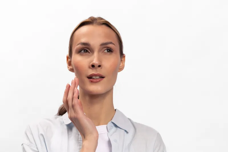 Thoughtful young woman with light skin and pulled back hair touches her face looking away on white background. Concept of skin care, beauty, natural look, and contemplation in studio lighting.