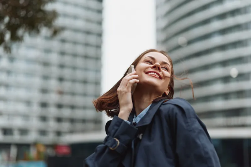 Confident woman in a navy jacket smiles while talking on the phone in a dynamic urban setting, tall buildings behind, candid street portrait. Bright city mood.