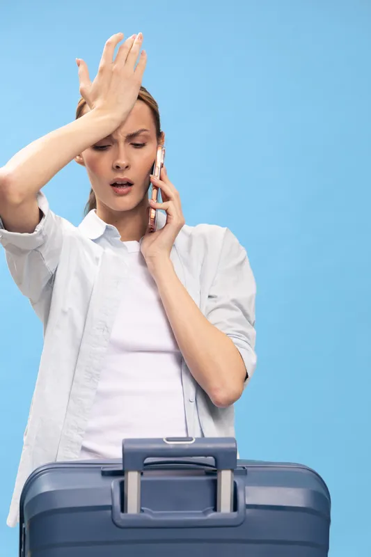 Stressed young woman talking on phone with frustrated expression, holding hand to forehead, standing behind blue suitcase on light blue background. Concept of travel problems and anxiety.