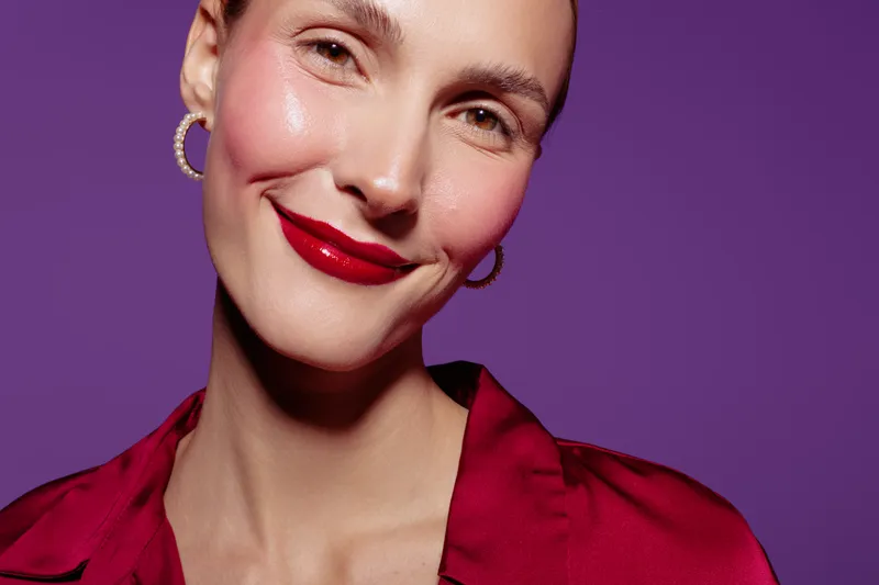 Close-up portrait of a smiling woman with red lipstick and hoop earrings wearing a red satin blouse against purple background. Concept of beauty, makeup, fashion, and confidence.