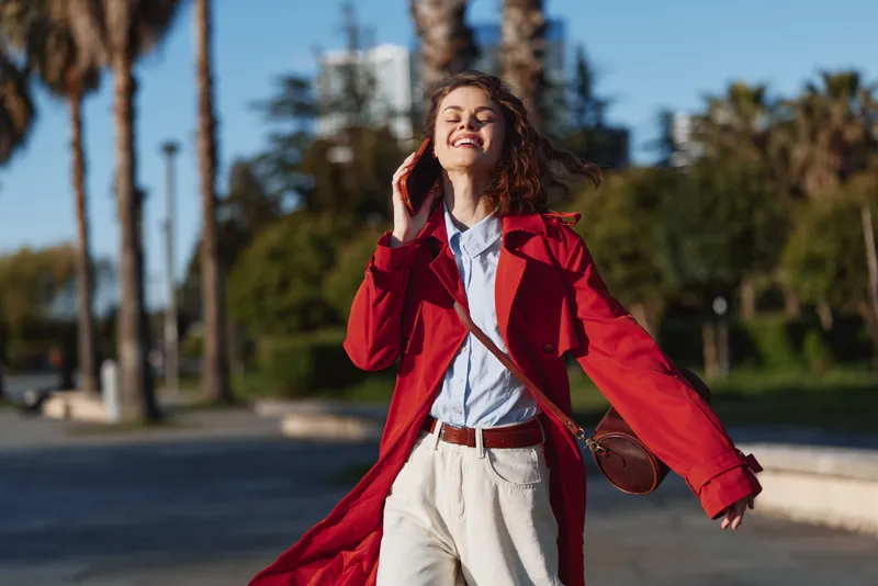 A happy young woman in a red coat talks on a smartphone outdoors on a sunny day, enjoying the fresh air and urban surroundings with palm trees in the background.