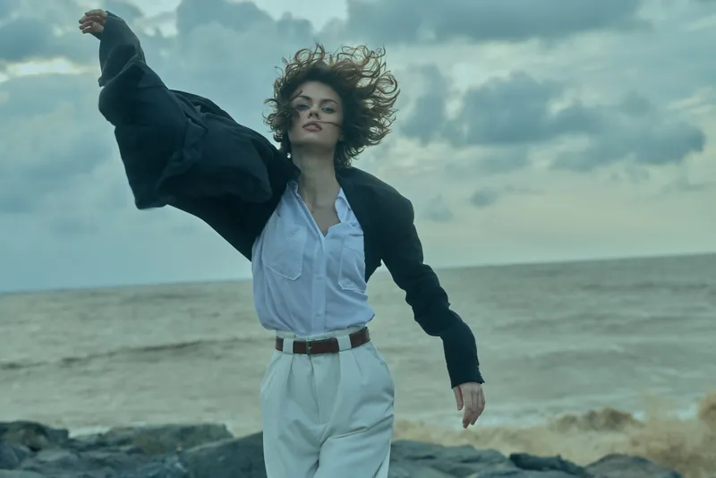 Woman on a windy coastline in a blazer and white pants, mid motion with sea breeze, bold confidence in a maritime fashion moment