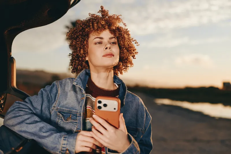 Woman selfie smartphone denim jacket curly hair sunset road travel lifestyle outdoors, female holds smartphone by car window, denim casual outfit, golden hour sunset, traveling, calm smile, portrait