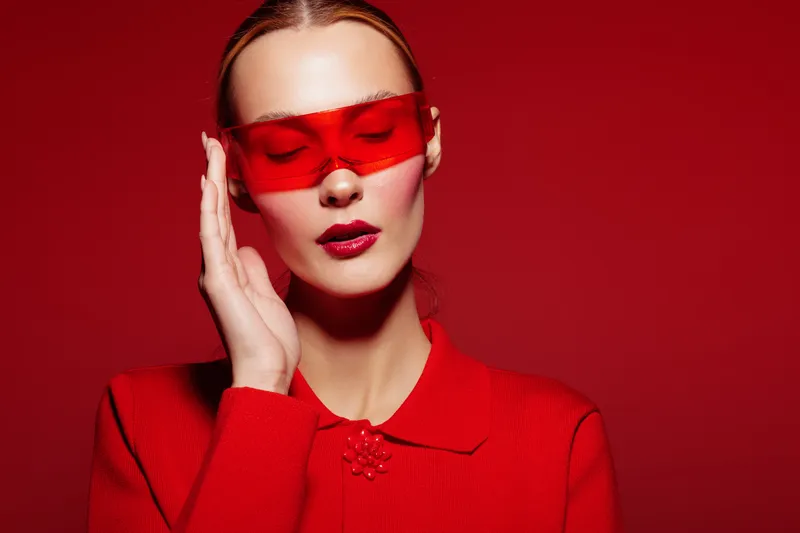 Stylish woman in red sweater and red sunglasses poses with closed eyes on red background. Fashion portrait with monochrome color scheme and modern accessories concept.