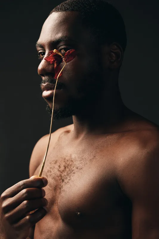 Creative conceptual portrait of African man holding dried flower near face with dark background, artistic visual style, bare chest, thoughtful expression, studio lighting, closeup profile.
