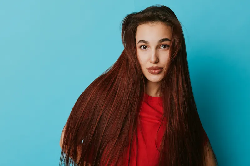 Young woman with long straight hair posing in red shirt against blue background. Portrait of female model with natural makeup and confident expression looking at camera.
