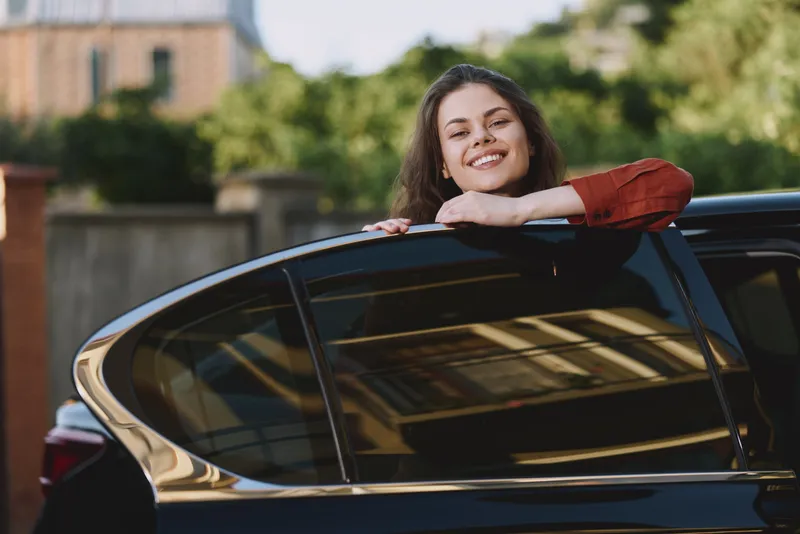 A cheerful young woman leans on a parked car, smiling at the camera in a sunlit street. A candid lifestyle portrait capturing confidence, happiness, and everyday moments